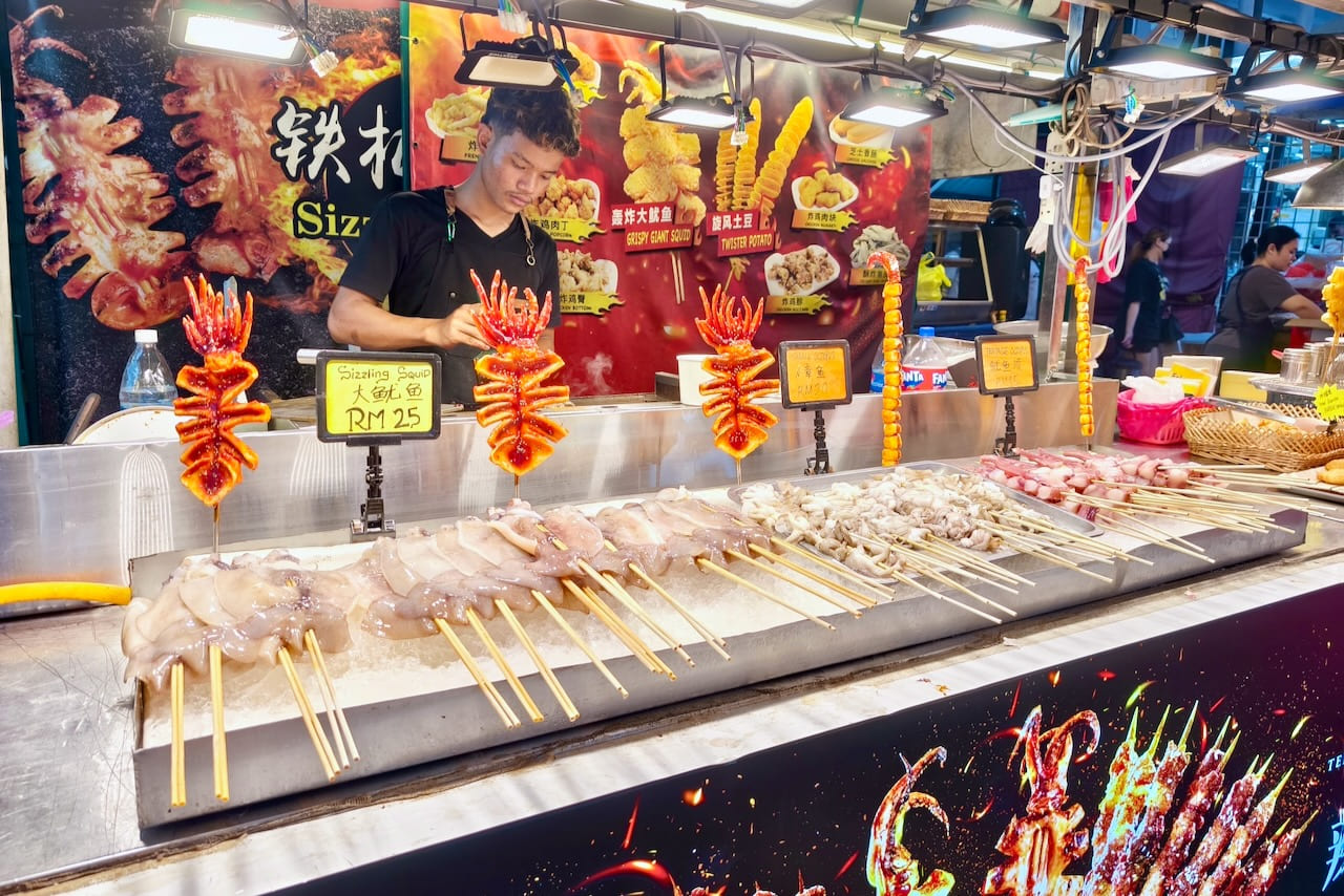 A man at a Malaysian street food stall grilling squid and other seafood on a hot grill.